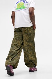 Person wearing a white T-shirt with a green snail graphic, loose green camouflage pants, and purple sneakers, standing with hands in pockets.
