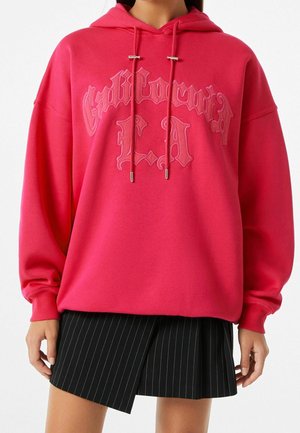 Donna che indossa un vivace hoodie rosa con testo gotico ricamato e una mini gonna plissettata nera a righe, su sfondo bianco.