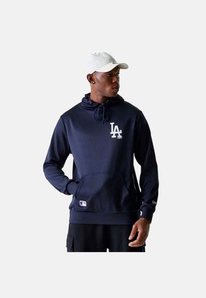 Mann trägt dunkelblauen LA-Logo-Hoodie und weiße Mütze, schaut zur Seite mit einer Hand in der Tasche vor schlichten Hintergrund.