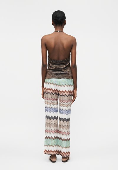 Γυναίκα με σκουρόχρωμο δέρμα και μαλλιά σε κότσο φοράει ένα καφέ τοπ χωρίς πλάτη και φαρδιά παντελόνια με πολύχρωμο ζιγκ ζαγκ μοτίβο, κοιτάζοντας προς τα πίσω.