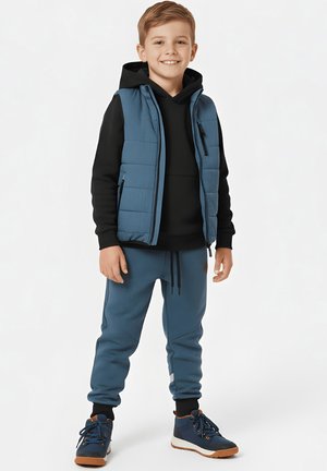Uśmiechający się chłopiec w czarnej bluzie z kapturem, niebieskiej pikowanej kamizelce, niebieskich spodniach dresowych i granatowych tenisówkach, stojący na jednolitym tle.