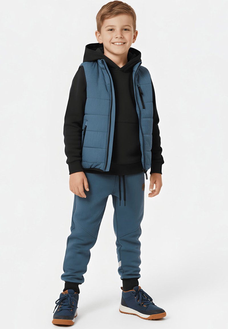 Uśmiechający się chłopiec w czarnej bluzie z kapturem, niebieskiej pikowanej kamizelce, niebieskich spodniach dresowych i granatowych tenisówkach, stojący na jednolitym tle.
