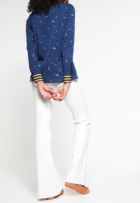 Mörkblå långärmad blus med ett litet guldmönster, med randiga manschetter. Kombinerad med vita utsvängda byxor och bruna skor.