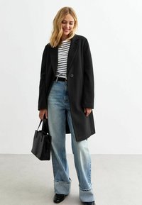 Manteau long noir avec un bouton, porté sur un haut ray�é. Associé à un jean baggy bleu clair et un sac à main noir texturé.