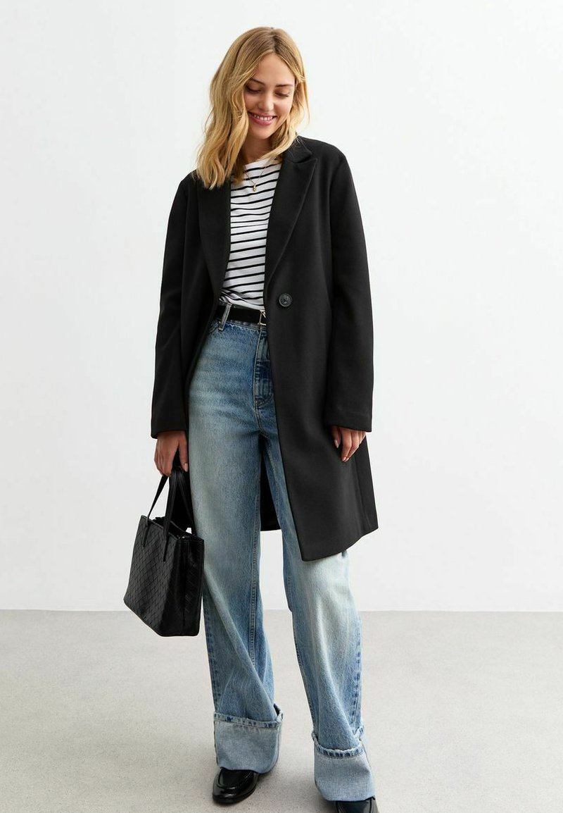 Manteau long noir avec un bouton, porté sur un haut rayé. Associé à un jean baggy bleu clair et un sac à main noir texturé.