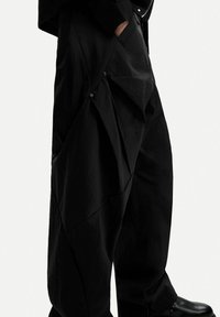 Black pants with a textured finish, featuring an asymmetric design, snap-button accents, and side pockets. Material appears lightweight.