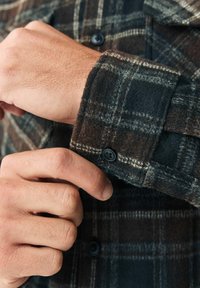 Hnědá a černá kostkovaná flanelová košile s knoflíkovým manžetou a texturovanou tkaninou. Záběr na ruku upravující rukáv.
