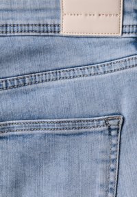 Jeans de mezclilla azul claro con una etiqueta de parche en la parte posterior con texto embosado, líneas de bolsillo curvadas y detalles de costura visibles.