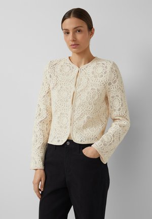 Kobieta nosząca kremową, kwiatową koronkową kurtkę z długim rękawem na jasnej bluzce i czarnych spodniach, stojąca z jedną ręką w kieszeni.