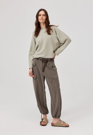 Kobieta stoi, ubrana w beżową, oversize bluzę, ciemnobrązowe spodnie typu cargo z paskiem i beżowe sandały na prostym jasnym tle.