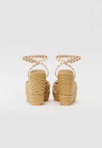 Sandalias de cuña con tacones de yute texturizados y diseño de correas cruzadas, con remaches metálicos en una parte superior de cuero sintético de color claro.