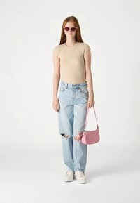 Νεαρή γυναίκα φορώντας μπεζ εφαρμοστό μπλουζάκι, σκισμένα ανοιχτόχρωμα τζιν, λευκά αθλητικά παπούτσια, ροζ γυαλιά ηλίου και κρατώντας ροζ τσάντα στο πλάι.