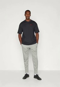 T-shirt nera a maniche corte con logo rosso, abbinata a pantaloni della tuta grigi chiari. Il modello indossa scarpe da ginnastica nere con calze bianche.