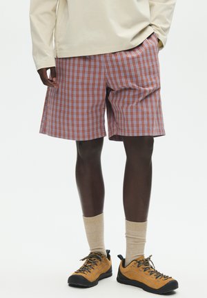 Mann trägt beige Langarmshirt, lila-orange karierte Shorts, beige Socken und hellbraune Wanderschuhe vor weißem Hintergrund.