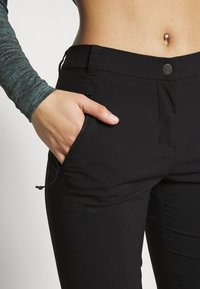 Black outdoor pants made of stretch fabric with a slim fit, featuring a zipper pocket and a button closure. Logo visible on the side.