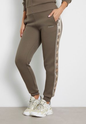 Kobieta w oliwkowych spodniach dresowych Guess z beżowym paskiem z logo po bokach, w białych masywnych trampkach, stojąca z jedną ręką w kieszeni.
