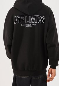 Sudadera negra con un gran diseño de texto blanco en la parte posterior que dice "ÁREA PELIGROSA FUERA DE LÍMITES" y "BERLÍN 2001". Cuenta con un bolsillo canguro.