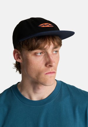 Młody mężczyzna z piegami, noszący czarno-granatową czapkę z pomarańczowym logo, patrzący w bok, ubrany w niebieską koszulkę na jednolitym tle.