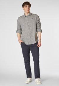 Camicia button-up grigia con una leggera trama, un piccolo logo sul petto, abbinata a pantaloni blu navy e sneakers bianche.