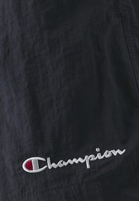 Material textil negru cu o finisare texturată are un logo brodat alb "Champion" accentuat cu un detaliu roșu.