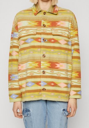 Osoba ubrana w zapinaną na guziki kurtkę z polaru z wielokolorowymi, poziomymi wzorami plemiennymi oraz niebieskie jeansy typu cargo.