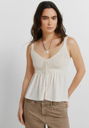 Kvinde iført en cremefarvet hæklede camisole-top og lysebrune jeans, stående med den ene hånd i lommen mod en ensfarvet baggrund.