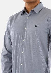 Camicia a maniche lunghe blu e bianca a righe con chiusura a bottoni e colletto; presenta un logo ricamato sul petto. Tessuto con una texture liscia.