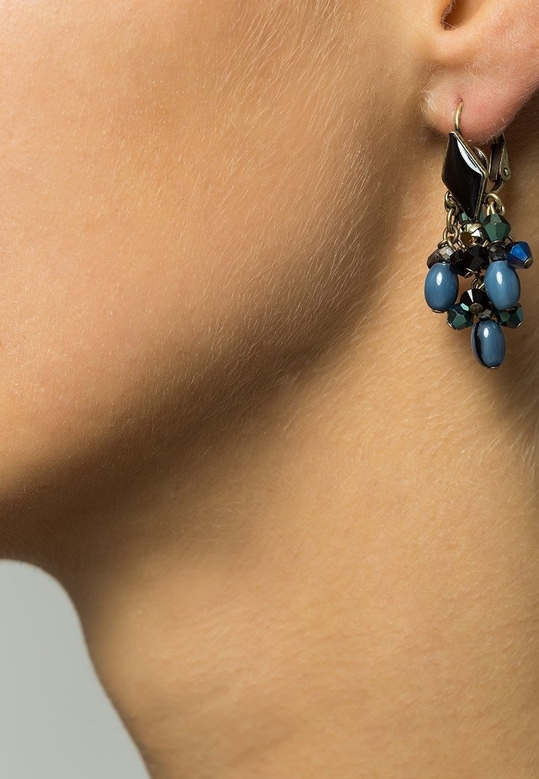 Gros plan sur un lobe d’oreille portant une boucle d’oreille dorée avec des perles noires, bleues et vertes regroupées dans un design pendentif.