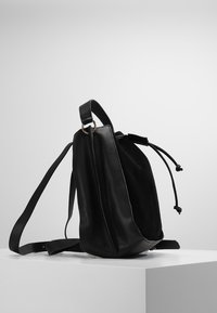 Musta nahka- ja mokkakankaisesta narulaukusta, jossa on muodollinen rakenne, säädettävät hihnat ja sileä pinta. Minimalistinen muotoilu.