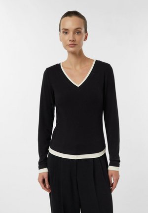 Woman wearing black long-sleeve V-neck top with white trim at neckline, cuffs, and hem, paired with black pleated pants, standing against white background.