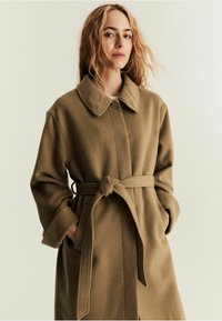 Langer Wollmischmantel in olivbraun mit Reverskragen, Knopfverschluss, tailliertem Gürtel und seitlichen Taschen. Weiche, strukturierte Oberfläche.