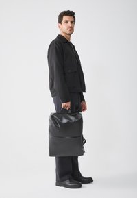 Bolso de hombro negro con textura suave y brillante, con asa superior y bolsillo con cremallera en la parte frontal, sostenido por una persona vestida de negro frente a un fondo neutro.