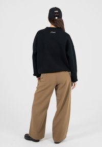 Eine Person trägt einen schwarzen Pullover und eine Kappe mit dem Text "Moderne Yuppies" sowie lockere braune Hosen. Sie steht mit dem Rücken zu einer schlichten Hintergrund.