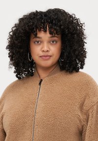 Veste bomber en polaire marron avec fermeture éclair sur le devant, col côté et texture douce. Le mannequin a des cheveux bouclés et porte des boucles d'oreilles créoles.