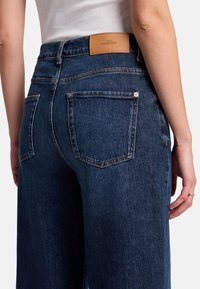 Mörkblå denimjeans med en lädermärke på baksidan av midjan, fem fickor och synliga sömnadsdetaljer.
