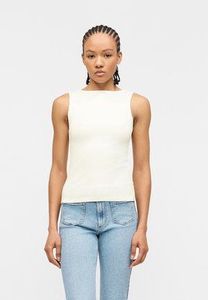 Νεαρό άτομο με πλεξούδες, φορώντας ένα λευκό αμάνικο τοπ και ανοιχτό μπλε τζιν ψηλόμεσο, στέκεται μπροστά σε ένα απλό φωτεινό φόντο.