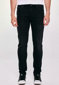 Czarne dżinsy slim-fit wykonane z denimu, z pięcioma kieszeniami, z zapięciem na guzik oraz delikatnym przetarciem dla nadania tekstury. Noszone z czarnymi trampkami.