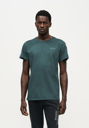 Un homme porte un tee-shirt Moschino bleu sarcelle avec des finitions aux épaules et un pantalon noir sur un fond blanc uni.