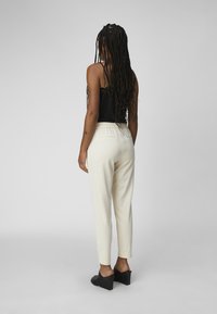 Femme aux longues tresses portant un haut sans manches noir, un pantalon couleur crème et des chaussures à talons noirs, se tenant dos à la camera.