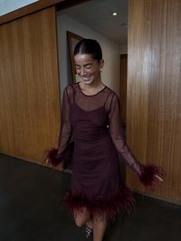 Vinröd klänning med genomskinliga långa ärmar och fjäderdetaljer vid muddar och fåll. Slimmad silhuett, slät textur, diskret design.