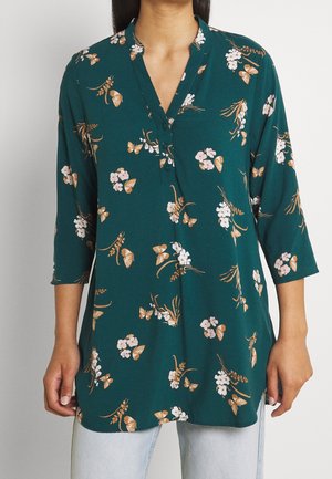 Bluzka z długim rękawem w kolorze morskim z zapinanym na guziki dekoltem w kształcie litery V, ozdobiona beżowymi i białymi wzorami kwiatów i motyli, noszona z jasnoniebieskimi dżinsami.