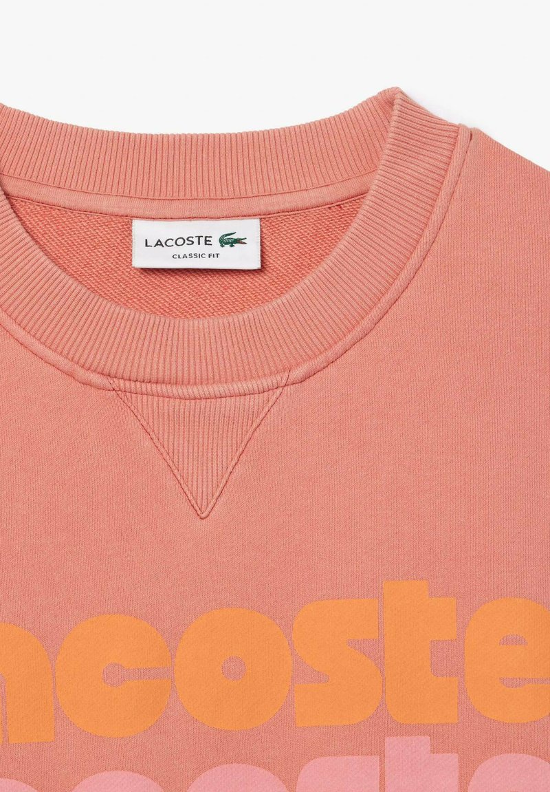 Felpa rosa chiaro con collarino a coste e dettaglio di cucitura triangolare. Presenta un testo arancione audace, etichettato "Lacoste Classic Fit."