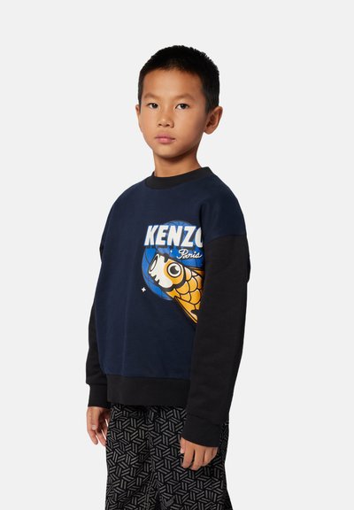 Sweatshirt bleu marine avec des manches longues noires, orné d'un graphique de poisson coloré et du texte "KENZO Paris". Associé à un pantalon noir à motifs.