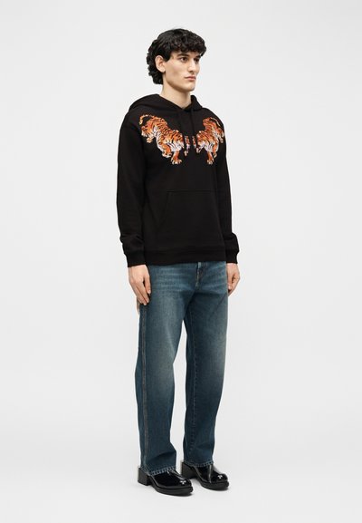 Jeune homme debout portant un sweat à capuche noir avec deux tigres orange imprimés sur la poitrine, un jean bleu et des chaussures noires brillantes sur un fond uni.