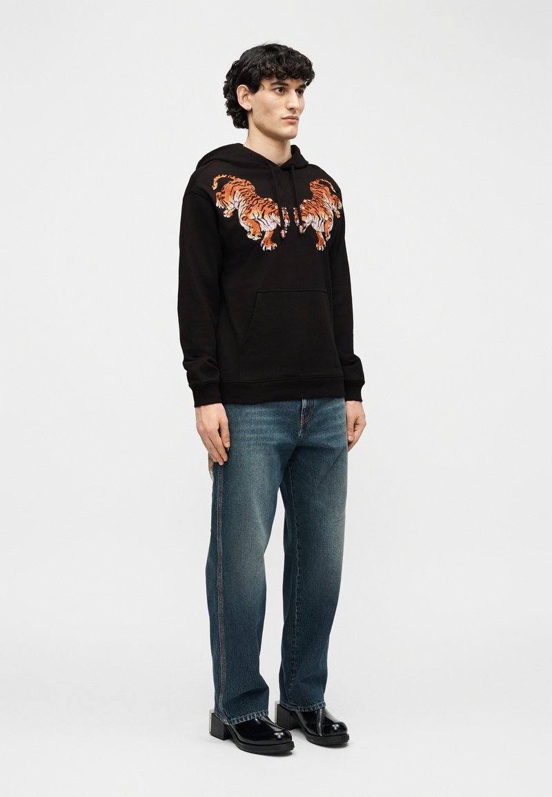 Giovane uomo in piedi che indossa una felpa nera con due tigri arancioni stampate sul petto, jeans blu e scarpe nere lucide su sfondo semplice.