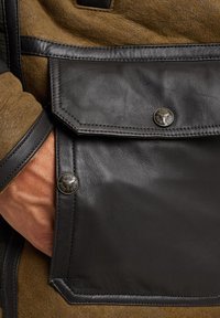 Bruine suede jack met een zwarte leren zak. Heeft een geometrische flap en twee bronzen knopen met reliëfdetails. Soepele textuur.