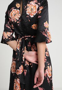 Černý květinový kimono s velkými oranžovými a růžovými květinami, měkká saténová textura, zvonové rukávy a vázací pás, s vzorovaným pozadím.