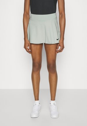 Osoba nosząca jasnoszarą sportową spódnicę Nike, czarny top, białe skarpetki i białe trampki, stojąca na jednolitym tle.