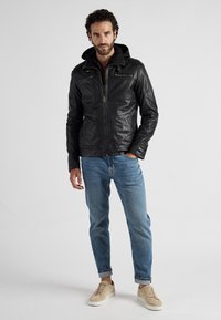 Zwart leren jack met een capuchon, met rits aan de voorkant, twee borstzakken en zijzakken. Draagt met lichtblauwe jeans en beige sneakers.