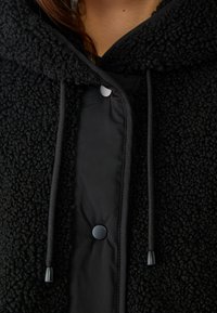 Schwarze Fleecejacke mit einer strukturierten, flauschigen Außenseite, die ein glattes Nylonfutter, Kordelzüge und Druckknopfverschlüsse aufweist.